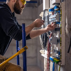 A male electrician works in a switchboard with an electrical connecting cable, connects the equipment with tools.